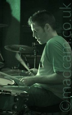 A man sitting behind a drum kit, with a drum stick in each hand that are blurred slightly as they move. He has short, slightly spiky hair, is wearing a light-coloured t-shirt and dark trousers, and is sitting facing to the left. The whole frame has a green tint.