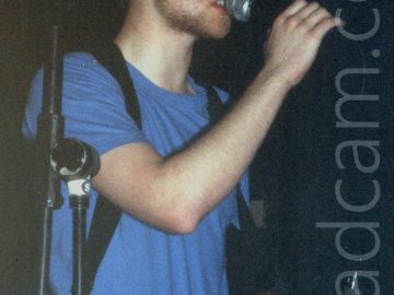 A man with short dark hair and short beard, wearing a blue t-shirt, and with a bass guitar attached to a black strap slung over both shoulders. He is holding on to a microphone on a stand with his right hand, as he sings in to it. Black walls and ceiling fill the rest of the frame.