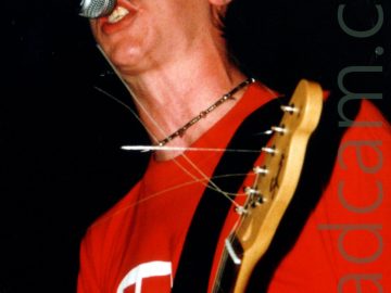 A man with a blue baseball cap over short dark hair, wearing a red t-shirt with a white, black, and red logo on the front, holding the head of an electric guitar in his left hand and singing into a microphone on a stand in front of him. Black walls and ceiling fill the background.