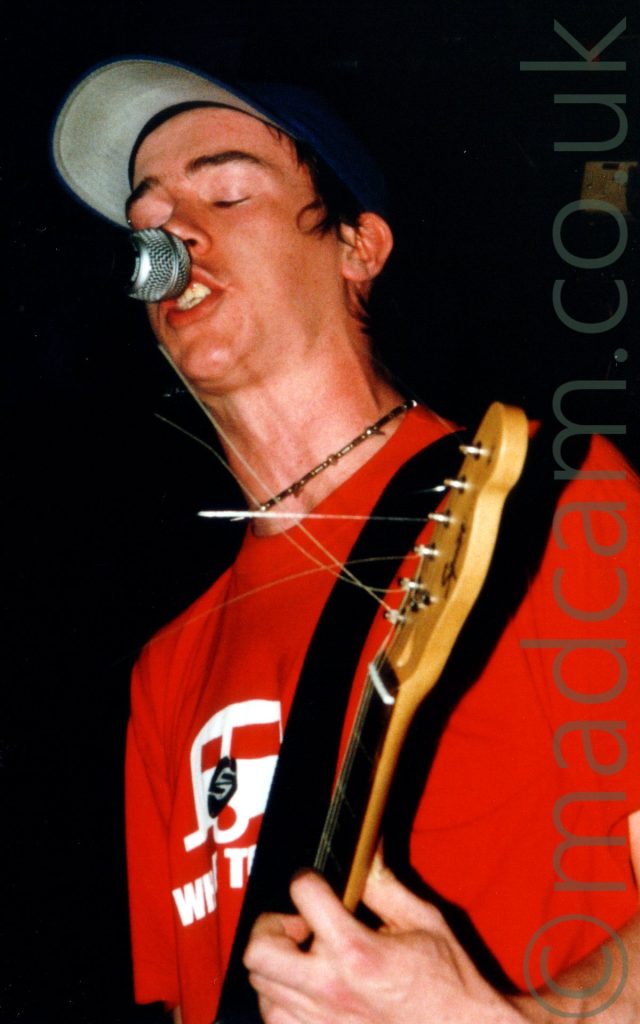 A man with a blue baseball cap over short dark hair, wearing a red t-shirt with a white, black, and red logo on the front, holding the head of an electric guitar in his left hand and singing into a microphone on a stand in front of him. Black walls and ceiling fill the background.