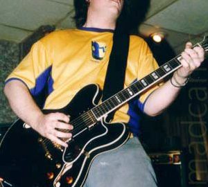 A man with shaggy dark hair, wearing a yellow t-shirt with blue sides and blue piping on the neck and sleeves, and a blue and white face on the front, as well as grey trousers. He is playing a black electric guitar, attached to a wide black strap over his left shoulder. Mottled green wallpaper covers the wall on the left of the frame, with white tiles on the suspended ceiling above him.