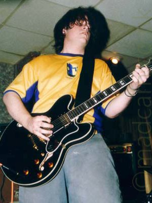A man with shaggy dark hair, wearing a yellow t-shirt with blue sides and blue piping on the neck and sleeves, and a blue and white face on the front, as well as grey trousers. He is playing a black electric guitar, attached to a wide black strap over his left shoulder. Mottled green wallpaper covers the wall on the left of the frame, with white tiles on the suspended ceiling above him.