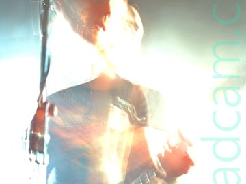 Long exposure shot of a man with short dark hair and wearing a black t-shirt with a brown image filling the chest, and a blue pair of jeans, playing a red electric guitar. The long exposure and the bright stage lights have washed out much of the stage area, while a snap zoom and his movement have left his head dis-jointed from the rest of his body. A microphone on a stand in front of him reflects the stage lights. Black walls and ceiling complete the image.