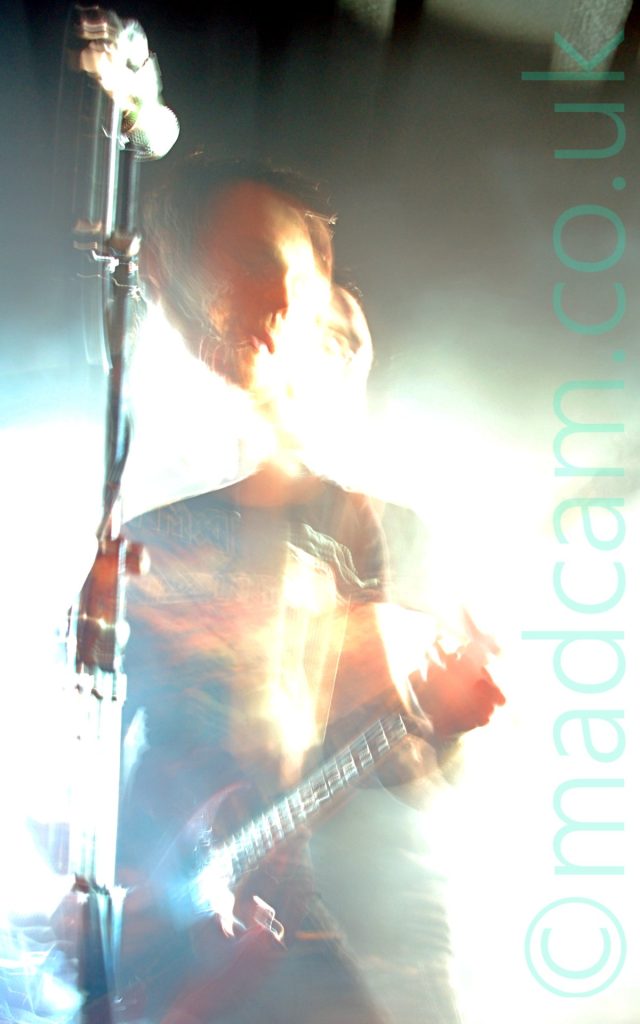 Long exposure shot of a man with short dark hair and wearing a black t-shirt with a brown image filling the chest, and a blue pair of jeans, playing a red electric guitar. The long exposure and the bright stage lights have washed out much of the stage area, while a snap zoom and his movement have left his head dis-jointed from the rest of his body. A microphone on a stand in front of him reflects the stage lights. Black walls and ceiling complete the image.