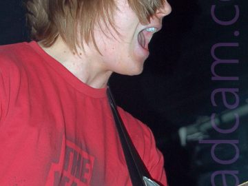 A man with shaggy, mid-length brown hair that covers most of his face, wearing a red t-shirt with a grey textual logo, playing a red electric guitar attached to a black strap slung over his left shoulder. His mouth is open as if he is singing, although there is no microphone in sight. Black walls and ceiling fill the rest of the frame.