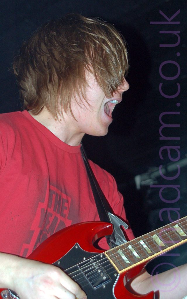 A man with shaggy, mid-length brown hair that covers most of his face, wearing a red t-shirt with a grey textual logo, playing a red electric guitar attached to a black strap slung over his left shoulder. His mouth is open as if he is singing, although there is no microphone in sight. Black walls and ceiling fill the rest of the frame.