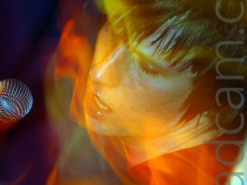 A man with shaggy dark hair, wearing a dark t-shirt, with a light-coloured guitar strap over his left shoulder as he leans backwards towards the camera, with a microphone coming into the frame from the left, beside his face. A long exposure combined with his movement and the stage lights leaves light trails and secondary images streaks of light across the frame.