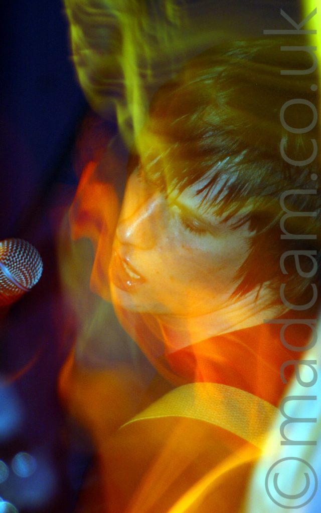 A man with shaggy dark hair, wearing a dark t-shirt, with a light-coloured guitar strap over his left shoulder as he leans backwards towards the camera, with a microphone coming into the frame from the left, beside his face. A long exposure combined with his movement and the stage lights leaves light trails and secondary images streaks of light across the frame.