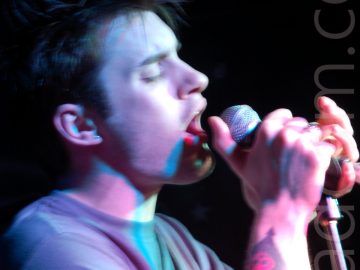 A man with short dark hair singing in to a microphone clasped in both hands, with his eyes shut. He has spiky brown hair, and a tattoo of an black love heart overlaid with a red skull on his right forearm. A long exposure and purple stage lights leaves some artifacts and streaks of light around his hands and face. Black walls fill the background.