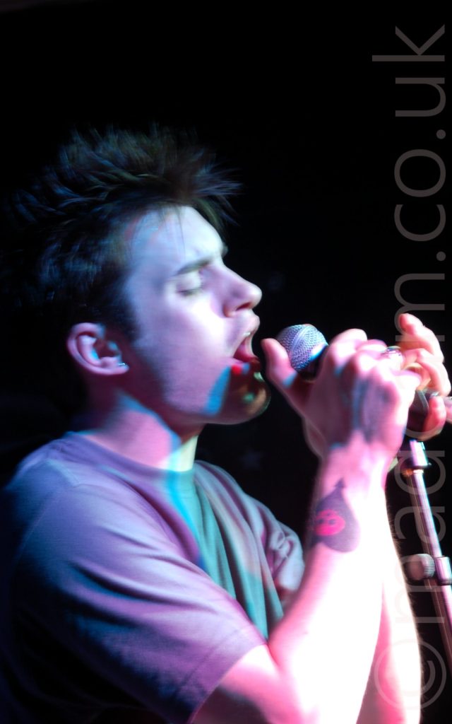 A man with short dark hair singing in to a microphone clasped in both hands, with his eyes shut. He has spiky brown hair, and a tattoo of an black love heart overlaid with a red skull on his right forearm. A long exposure and purple stage lights leaves some artifacts and streaks of light around his hands and face. Black walls fill the background.