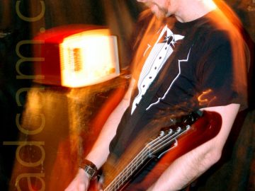 A Man with short dark hair, wearing a black and white t-shirt that resembles a tuxedo with a white shirt and bow tie, standing facing the left of the frame while playing a brown bass guitar. A flash from the camera has frozen him in place, while a long exposure, his movements, and the muted stage lights have left yellow and orange secondary images around him. An amplifier with a weird looking box on top is in the bottom left corner, with textiles draped over the walls in the background.