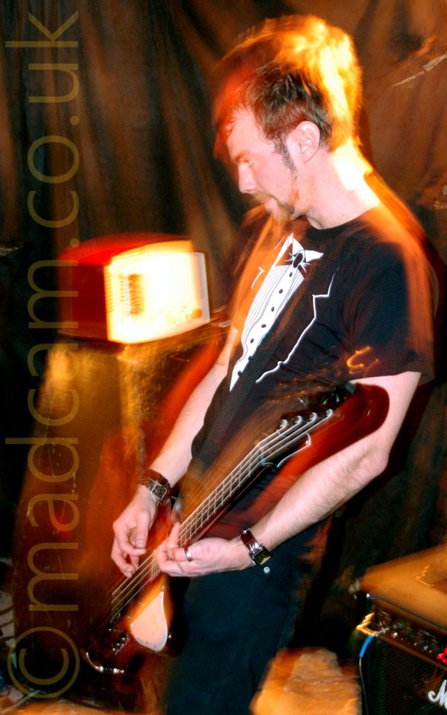 A Man with short dark hair, wearing a black and white t-shirt that resembles a tuxedo with a white shirt and bow tie, standing facing the left of the frame while playing a brown bass guitar. A flash from the camera has frozen him in place, while a long exposure, his movements, and the muted stage lights have left yellow and orange secondary images around him. An amplifier with a weird looking box on top is in the bottom left corner, with textiles draped over the walls in the background.