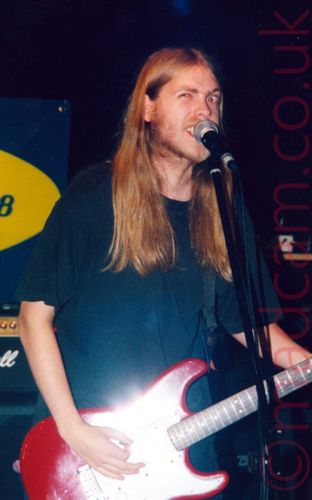 A man with long, blond hair, wearing a black t-shirt and playing a red electric guitar, scowling at the camera from behind a microphone on a stand. Black walls fill the background, with part of a dark blue poster with a yellow oval in the middle, on the left of the frame, above a stack of amplifiers.