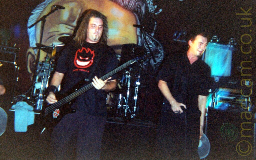 A rather grainy shot of 2 men on stage, performing as part of a band. On the left is a man wearing a black t-shirt with a red logo of a grinning face in the middle of the chest, and dark blue jeans, playing a black bass guitar attached to a black strap that is slung over his left shoulder. He has shoulder-length dreadlocks, and a black wristband around his right wrist. On the right is a man with short dark hair, wearing a black shirt and dark blue jeans, holding a microphone down by his waist in his right hand, looking out over the audience. A drum kit sits on an elevated podium behind them, with rack of amplifiers on the right of the frame. A large poster of a face is hanging on the wall at the back, partially covered by the drum kit.