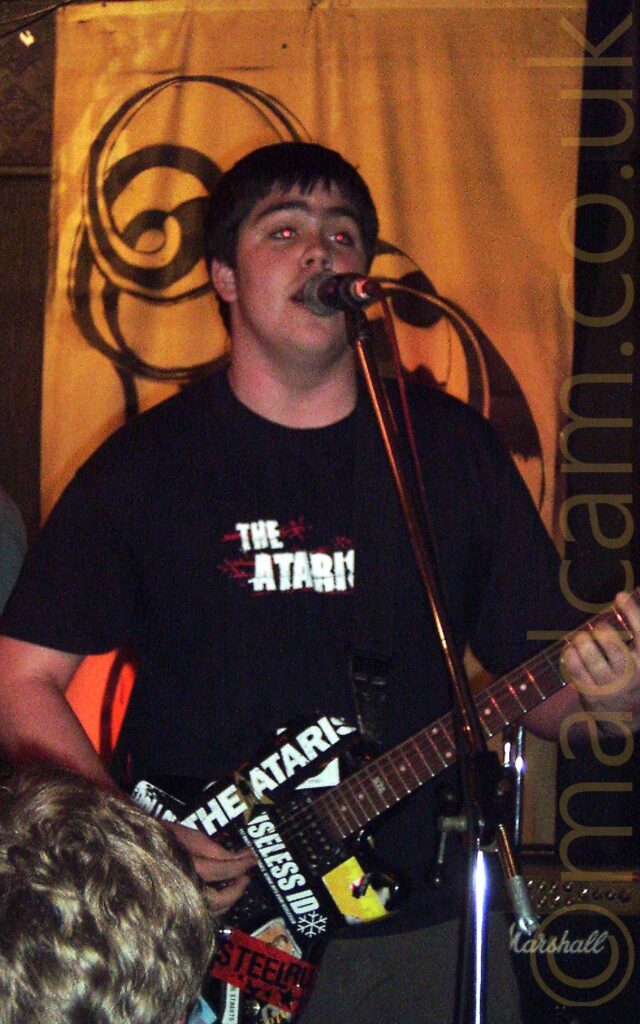 A man with short dark hair, wearing a black t-shirt with white text "The Ataris" and playing a black electric guitar festooned with white stickers (some reading "The Ataris" and "Useless ID" while singing into a microphone on a stand directly in front of him. The back of the head of someone in the crowd can be seen in the bottom right corner. A golden drape with an ornate circle-based black logo hangs on the back of the wall at the back of the stage.