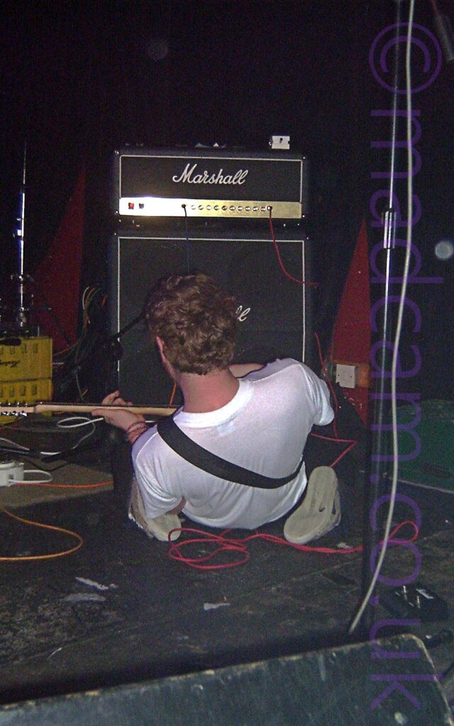 A man with his back to the camera, on his knees and lying backwards, the soles of his footwear visible underneath him. He has wavy brown hair and is wearing a white t-shirt, with a guitar strap over his left shoulder attached to a guitar in his hands. A black guitar amp with a silver "Marshall" logo is in front of him at the back of the stage. Red walls covered in black curtains fill the background.