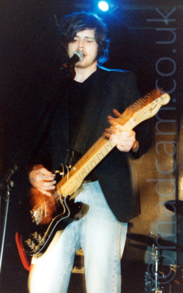 A man with wavy dark hair, wearing a black suit jacket over a black t-shirt and a blue pair of jeans, playing a black electric guitar, and singing into a microphone held on a stand in front of him. A flash has frozen him into place, a long exposure showing yellow secondary images where his arms and guitar have moved. A drumkit can be seen in the bottom right corner behind him, with a brown wall beyond that, the wall slowly turning black as it merges into the roof. A single blue light shines over his head.