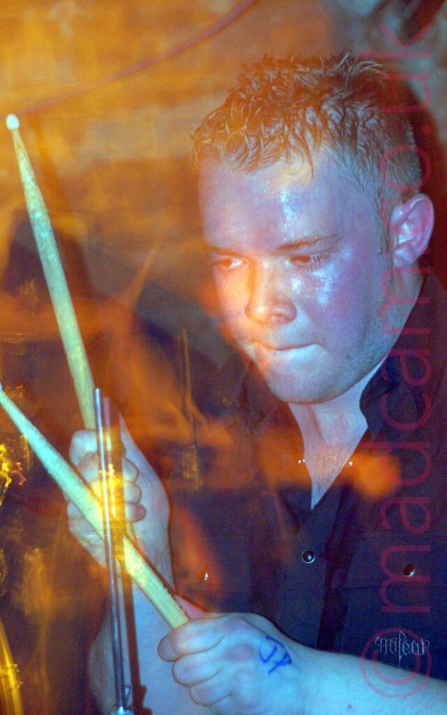 Close-up of a man with short, slightly spikey hair, wearing a black shirt, holding a pair of drumsticks raised in the air. A camera flash has frozen him in place, while a long exposure paints the frame with yellow lights.