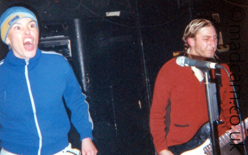 2 people performing on a stage as part of a rock band. On the left is a woman wearing a close-fitting blue hat that almost matches her blue tracksuit top, snarling at the crowd. On the right is a man with long brown hair tied back in pigtails and sporting some almost impressive sideburns in his beard. He is wearing a red jumper over a white shirt, and is playing a large black bass guitar, attached to a black strap over his left shoulder. Black walls and ceiling fill the background.
