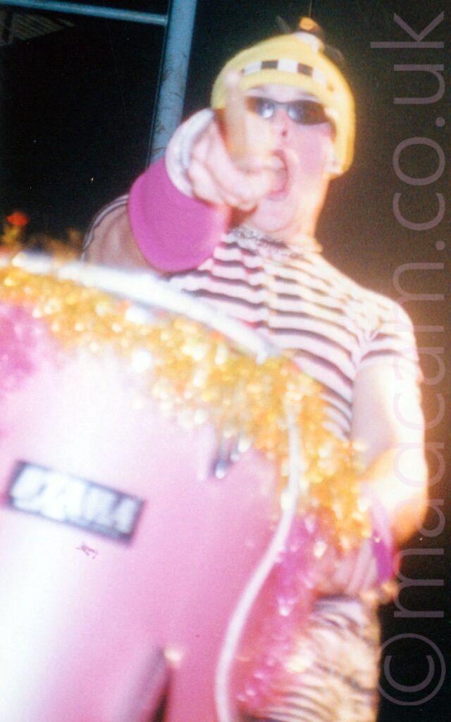 A man wearing a blue woollen hat, very dark sunglasses, and a stripey black and white top and trousers, as well as pink bands around his wrists, sitting behind a pink drum kit, and pointing the drum stick in his right hand at the camera, with his mouth agape. Black walls and ceiling fill the background.