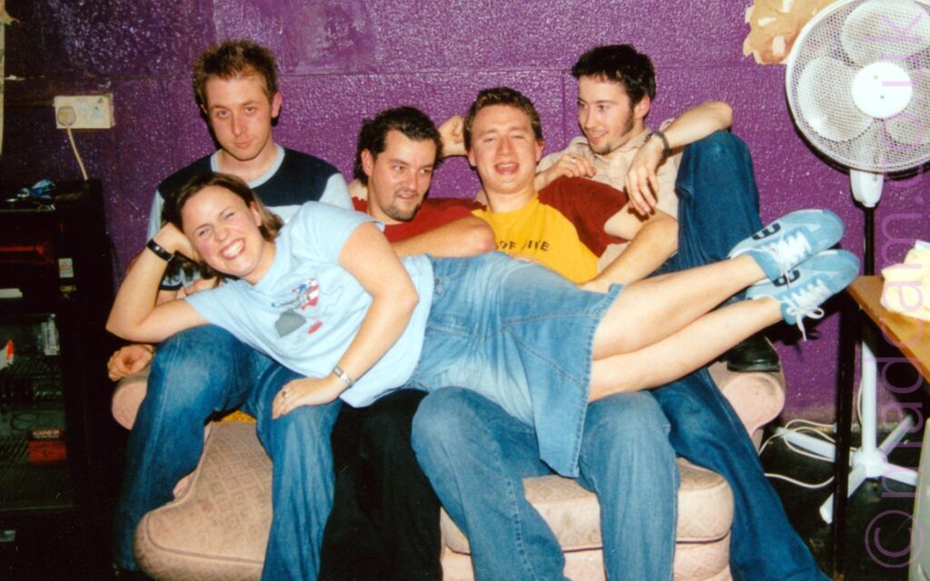 4 men with dark hair and wearing coloured t-shirts sitting on a rather battered low white sofa, with a blonde woman wearing a blue t-shirt, long denim skirt, and a massive mischievous grin on her face, draped decorously over their knees Sparkly purple paint covers the wall behind them, with a low, black drinks fridge in the background on the left, and a tall white fan and a brown table on the right.