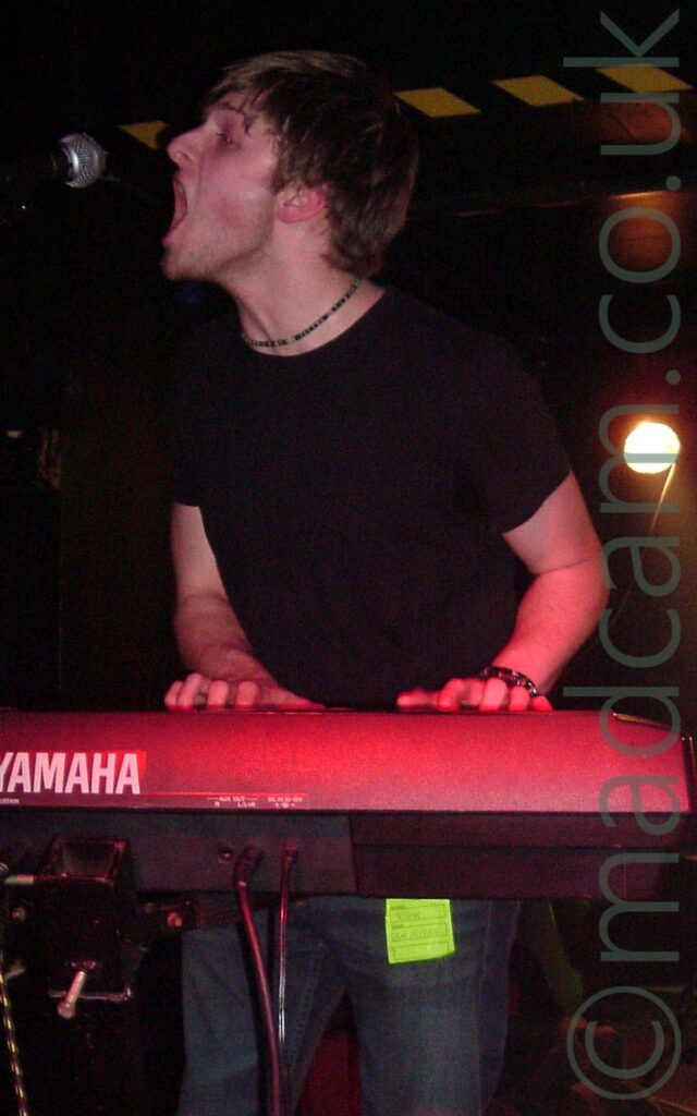 A man with short dark hair, wearing a black t-shirt and dark coloured trousers, playing a grey keyboard that appears to be tinted red from the stage lights, and singing (or possibly shouting) into a microphone. A yellow and black stripe runs across the black background wall behind his head. A yellow light shines almost directly into the camera on the right of the frame.