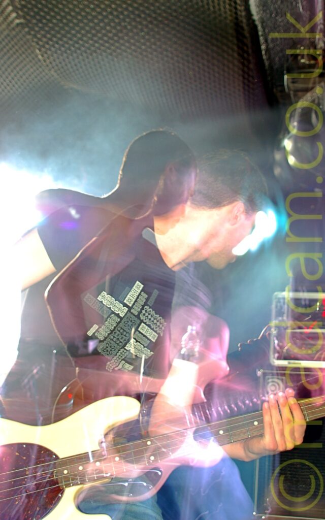 A man on a stage, with short dark hair, and wearing a black t-shirt with a creamy-green logo, and a blue pair of jeans, and playing a white bass guitar. A flash has frozen him in place, while a long exposure and slight zoom out allows movement, with 2 distinct images of the same person, with secondary images and red, blue, and white light auras around body parts and the walls in the background.