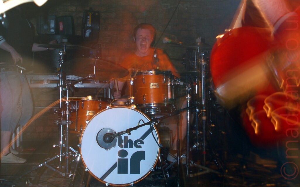 The drummer from a band sitting behind his kit at the back of a stage, and screaming for all he is worth as he plays. He is wearing an orange t-shirt, which pretty much matches the sides of his drums, and has short dark hair. The bass drum at the front has a white face, with a diagonal grey strip, overlaid with the grey text "The IR". Bright lights mounted on metal racks are shining down from the ceiling, in front of a brick wall. The head of a bass guitar is poking in to frame from the left, with the bottom of a red guitar on the right, with a large expanse of black floor between them.