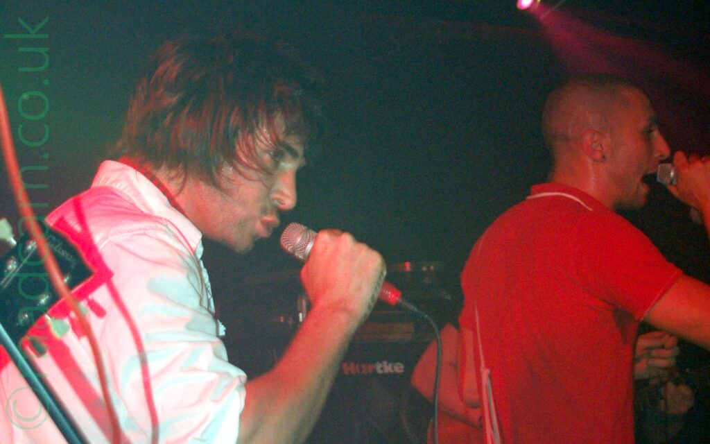 2 men, members of a band, performing on a stage, both fitting to the right. Nearest the camera, on the left of the frame, is a man with long, shaggy hair, wearing a white shirt, and singing into a microphone held in his right hand. On the right is a shaven-headed man wearing a red t-shirt with white piping, singing into a microphone in his right hand. A single red light in the top right is shining towards the front of the stage. Amplifiers line the black walls in the background.