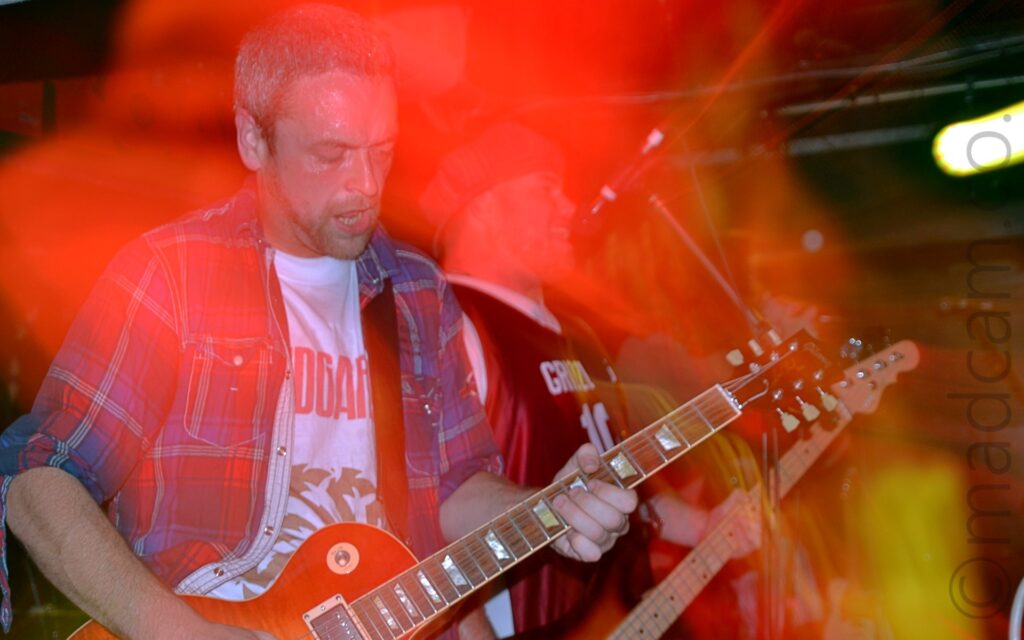 A man with short, close-cropped greying hair and beard, and wearing an open blue, red, and white checked shirt over a white t-shirt, playing an orange electric guitar. A camera flash has frozen him in place, while a long exposure and a slow zoom in has combined with the stage lights to paint the area around him with red and yellow light. In the background, a man wearing a blue cap and wearing a red and white basketball shirt and playing a bass guitar is singing into a microphone. Black walls mark the edges of the room, with a bright light smeared into a streak in the top right corner.