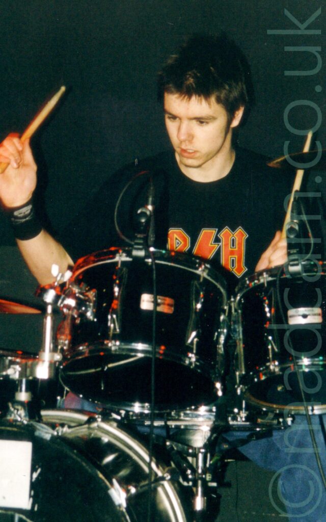 A man with short dark hair, wearing a black t-shirt with an indistinct red logo and a blue pair of jeans, sitting behind a black drumkit, with drumsticks held aloft. A black wall fills the background.