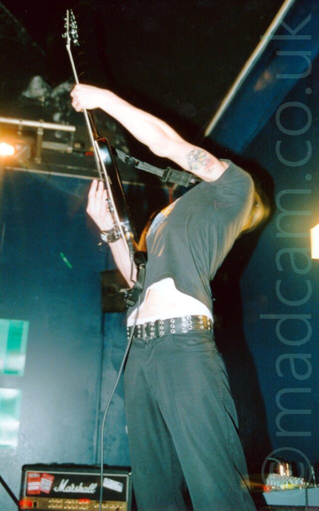 A low shot of a man playing a black electric guitar that is raised in the air as he leans backwards. He is wearing a black t-shirt and black trousers, the t-shirt rising to expose his belly. There is a leather strap on his right wrist, and a colourful tattoo on the back of his left bicep. Dark blue walls and black ceiling fill the background.