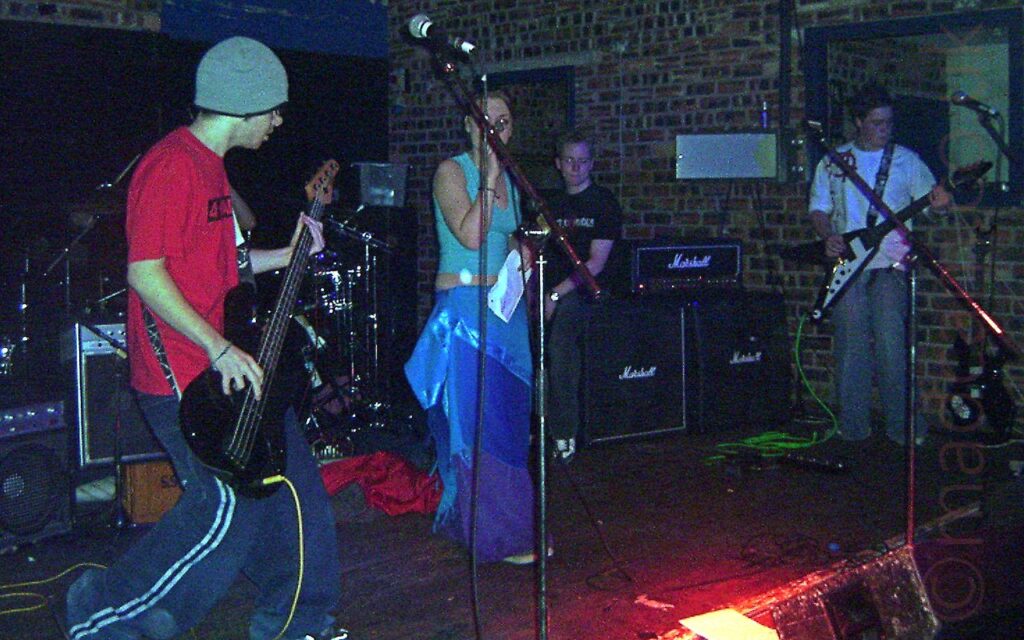 Somewhat grainy photo of several members of a band performing on a stage. On the left is a man with a grey woolen hat, and wearing a red t-shirt and dark blue baggy trousers with a double red stripe running down the leg, playing a black bass guitar. In the middle, partially hidden by a microphone stand, is a woman wearing a blue sleeveless top with part of her belly shown, as well as purple trousers with a blue sash tied around her waist, singing into a microphone held in her right hand.Behind her, a man with short, spiky hair and wearing a black t-shirt and jeans, is sitting on some black speakers, looking at the camera. On the right is a man with short dark hair, wearing a white t-shirt and blue jeans, playing a black flying v electric guitar. A brown brick wall fills most of the background.