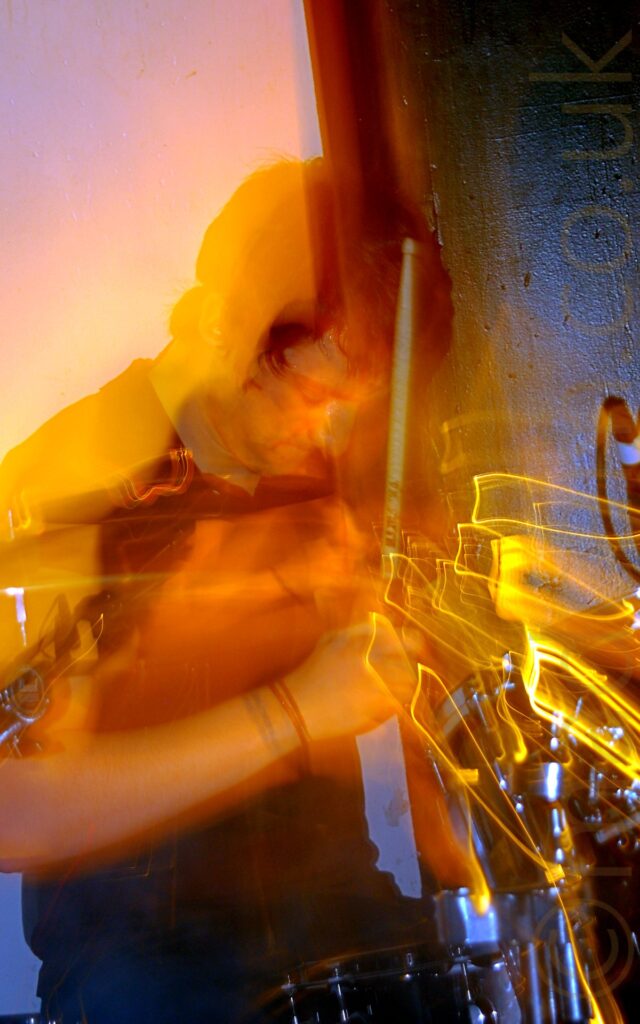 A man with short dark hair, wearing a dark coloured top, sitting behind a black drumkit, with a stick raised in the air in front of him, his head looking down. A flash has frozen him in place, while a long exposure and a snap zoom has created streaks of orange light and auras around any parts of him that have moved. A black wall on the right of the frame and a white wall on the left fill the background.