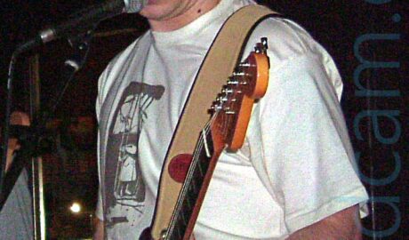 Slightly grainy photo of a man with short brown hair, wearing a white t-shirt with an indistinguishable black logo, playing a black electric guitar attached to a strap over his left shoulder, singing into a microphone while looking down at his instrument. Someone's left arm is just visible on the left of the frame. Black walls and ceiling fills the rest of the frame.