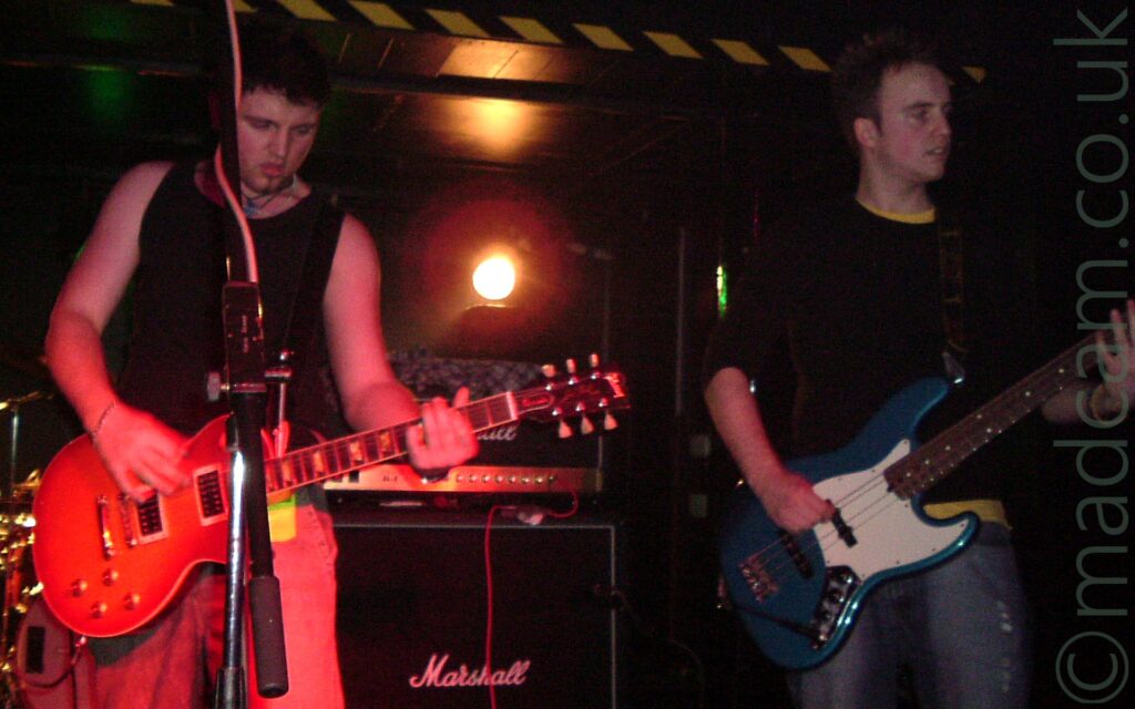 2 members of a band performing on a stage in a black room. On the left is a man with short dark hair, wearing a black vest and light-coloured jeans tinged red by onstage lighting, and playing an electric guitar. On the right is a man with short, spiky brown hair, wearing a dark top over a yellow t-shirt, and grey jeans, playing a blue, 4-string bass guitar. In the background, a stack of black speakers with white "Marshall" titles in in the bottom middle of the frame. A strip of tape with black and yellow diagonal stripes runs across the ceiling In the centre of the frame is a red light, giving off an almost HAL-9000 from the movie 2001 feel.
