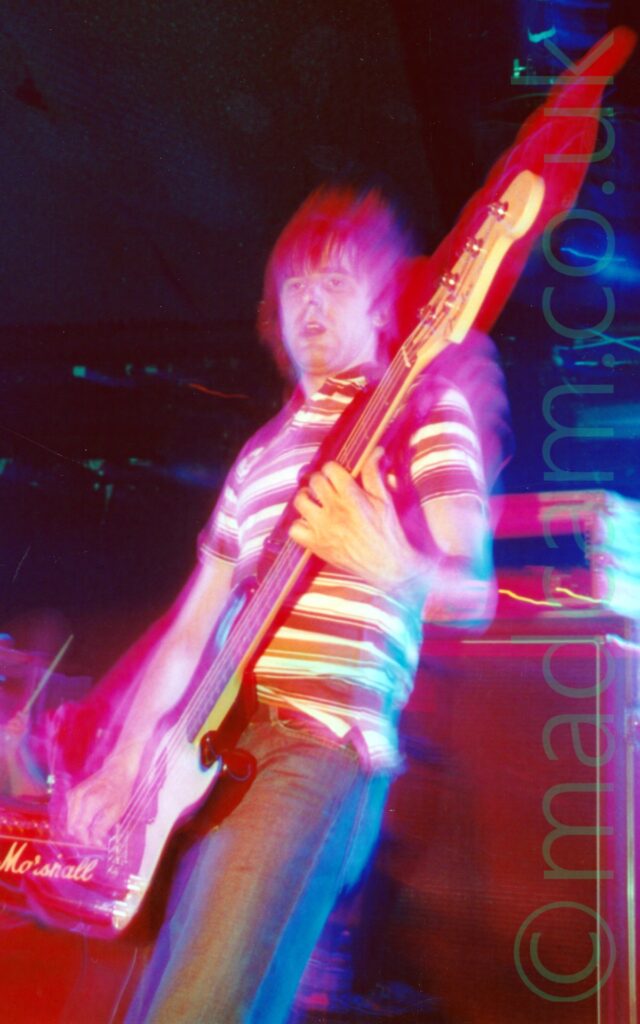 A man with short, dark hair swept down over his forehead, wearing a whit polo shirt with brown and orange bands around the body, and a blue pair of jeans, standing on a stage and playing a brown and white bass guitar. A flash has frozen him in time, while a long exposure, a slight zoom, his movement, and the stage lights have combined to paint the area around him in red. Amplifiers sit on the stage behind him, also tinted red, while the walls and ceiling at the back of the stage are black, with green speckles of reflected light on metal frames right at the back.