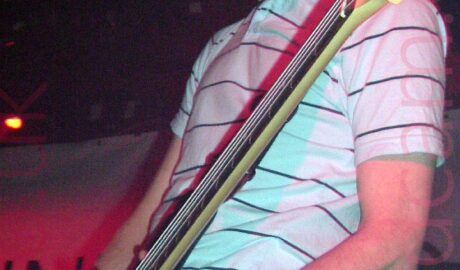 A man with mid-length dark hair, wearing a white polo shirt with thin black horizontal stripes and blue jeans, with a green and white label on the top of the left leg. He is playing a black electric bass guitar, and staring off into the distance to the left of the frame. A white sheet hangs on the black wall in the background, illuminated by a red light.