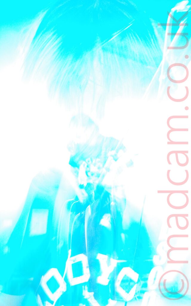 A massively over-exposed long exposure mixed with a snap zoom and an explosion of stage lights, leaving only a vague impression of what is actually there. It starts with a close-up of the head and upper body of a man with shoulder-length hair and wearing a dark t-shirt with a white text logo. His face and most of the background are blown out, massively over-exposed, along with the logo on the t-shirt, while dark areas of the image (his hair, t-shirt, and part of the ceiling behind him) are a light blue. A snap zoom out has a full length view of him, slightly hunched over, playing a guitar. Again, all details have been lost, blown out by the exposure, only blue shadows remaining.