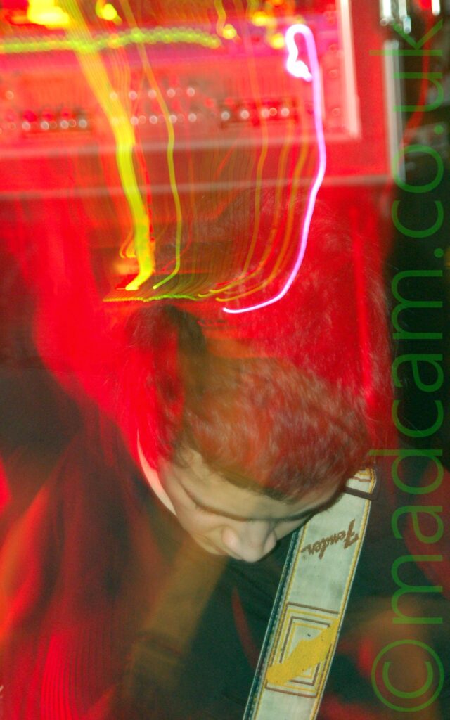 A man with short dark hair, and wearing a black top with a white Fender guitar strap over his left shoulder, hunched over as he plays his instrument. A camera flash has frozen him in place, while a long exposure, the stagelights, and his movements have left red, yellow, and white trails of light and secondary images as he headbangs his way through the photo.