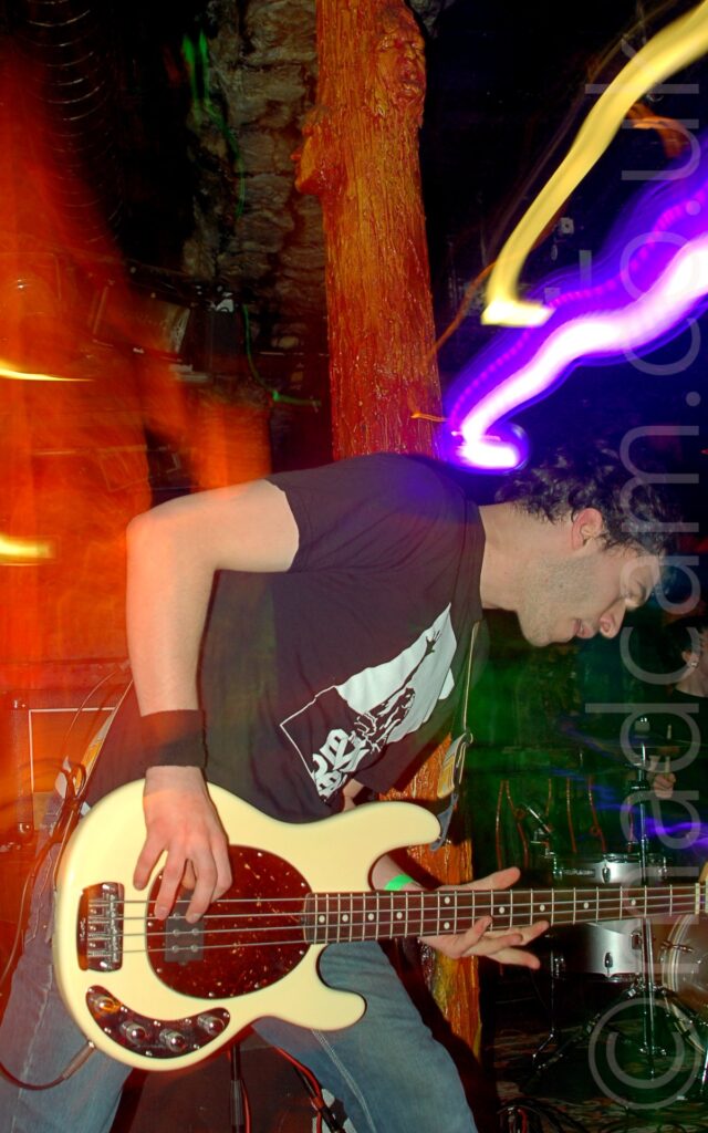 A man with short black hair, wearing a black t-shirt bearing a large, square, white and black logo, and a pair of blue jeans, playing a white, electric bass guitar. He is bent over, leaning to his left. A flash from the camera has frozen him in place, while a long exposure and a snap zoom has left an orange aura on the left of the frame, and streaks of yellow and purple light on the right. An orange pole with several faces on it, painted and glazed to look almost organic, stands behind him, while the bands drummer can be seen in the bottom right of the frame. Dark walls and ceiling fill the rest of the frame.