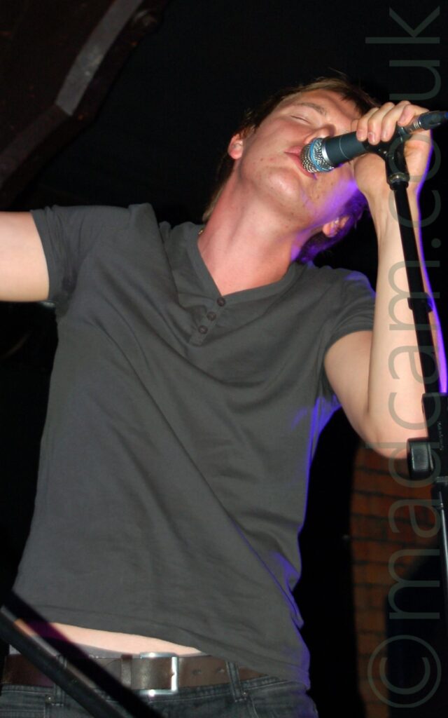 A man with short brown hair, wearing a green t-shirt and black jeans, leaning in to a microphone held on a stand in front of him, crooning in to the mic. Purple light is shining on his left side. Brown brick walls partially covered by black curtains are in the background, with black ceilings with ornate brown wooden roof supports filling the rest of the image.
