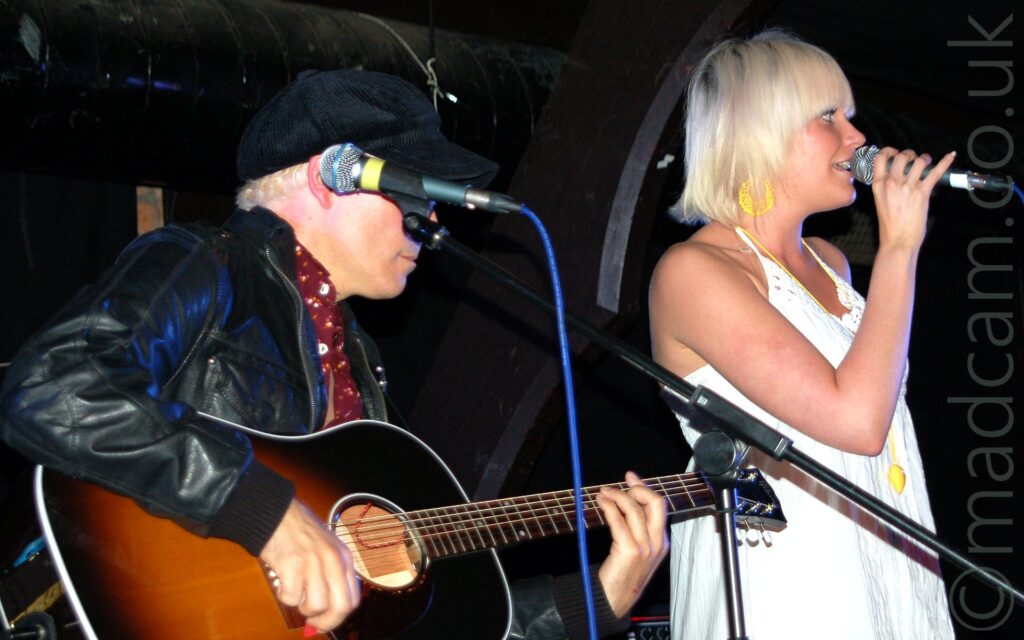 A woman with blonde hair standing on the right side of a stage, wearing a yellow sun dress, holding a microphone to her mouth as she sings, while looking off to the right of the frame. On the left is a man wearing a corduroy cap and a black leather jacket over a red shirt, playing a black acoustic guitar with a yellow sunburst, his face partly hidden by a microphone on a stand in the foreground. Black wooden beams and wide black metal tubes are against the black brick walls in the background.