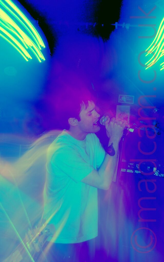 Long exposure of a man with dark hair, wearing a white t-shirt and a blue pair of jeans, holding a microphone in both hands as he sings. A long exposure shot, starting zoomed out then zooming in, with no flash. The result has clouds of blue and purple lights around his head, with streaks of green lights in both top corners, with secondary images of the singer behind him.
