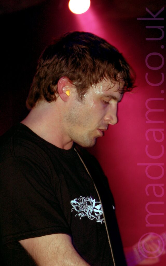 Side view of the head and shoulders of a man facing to the right, with short brown hair and wearing a black t-shirt with a white scrollwork logo in the middle of the chest. A black strap over his left shoulder suggests he has a guitar in his hands, which are out of frame. Black walls fill the background, with a red light in the top middle of the image flooding the background to the right with red light.