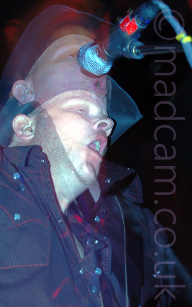 Long exposure of a man wearing a black stetson hat and a black shirt with red piping around the pockets. There are 2 images of him in the frame, the lowest is frozen in place because of a flash, then another as he moves while the shutter is open. In the upper image, he is singing into a microphone in front of him. Red light is painted around his head. The walls in the background are entirely black.