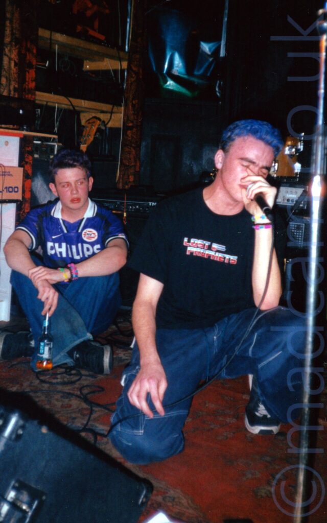 A man, with short blue hair, wearing a black t-shirt with a blue and red "Lost Prophets" logo on the nest, and a baggy pair of blue jeans crouched down on one knee on a tatty red carpet on a stage. He is holding a microphone to his mouth in his left hand, his right hand resting on his right knee. A black wedge monitor speaker is in the foreground in the bottom left corner, by his right knee. A young lad is sitting on the floor of the stage in the background on the left of the stage, with speakers and amps behind both of them, in front of black and red walls.