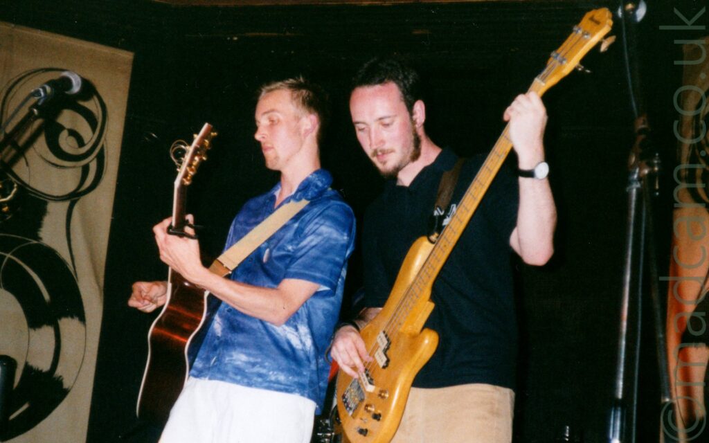 2 members of a band performing on stage. On the left is a man with short blond hair, wearing a blue, short sleeved shirt with white flecks, and white shorts, playing a brown acoustic guitar, with a tan guitar strap slung over his left shoulder. On the right is a man with short dark hair and a straggly beard, wearing a black polo shirt and light brown trousers, playing a pale-coloured bass guitar. Tan sheets of cloth with ornate black decorations cover sections of the black walls in the background.