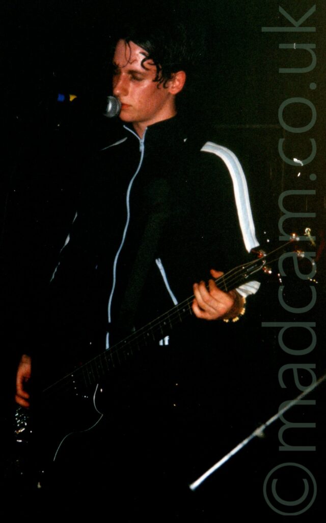 A man with straggly dark hair, and wearing a black jacket with thick white stripes on the sleeves and zips on the front and the pockets, playing a black bass guitar while singing in to a microphone on a stand, with his eyes shut. Black walls fill the background.