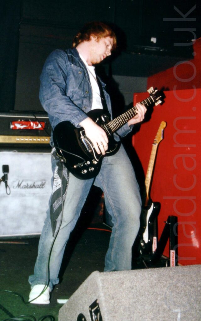 A man standing on a stage, with short ginger hair, wearing a denim jacket over a white t-shirt and faded blue jeans, and playing a black electric guitar while leaning back slightly. A grey felt-covered wedge monitor speaker is on the floor in front of him at the bottom right of the frame. Green carpet covers the rest of the foreground and part of the background, with a red half-height wall on the right, with a black and white electric guitar leaning against it. A large black amplifier with "Marshall" written on the front and a large red sticker with a pair of cartoon eyes on the top section is against the black walls at the back of the stage. Black walls and ceiling fill the rest of the image.